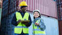 Man and woman, wearing high visibility jackets and hard hats, laughing in front of stacked shipping containers.