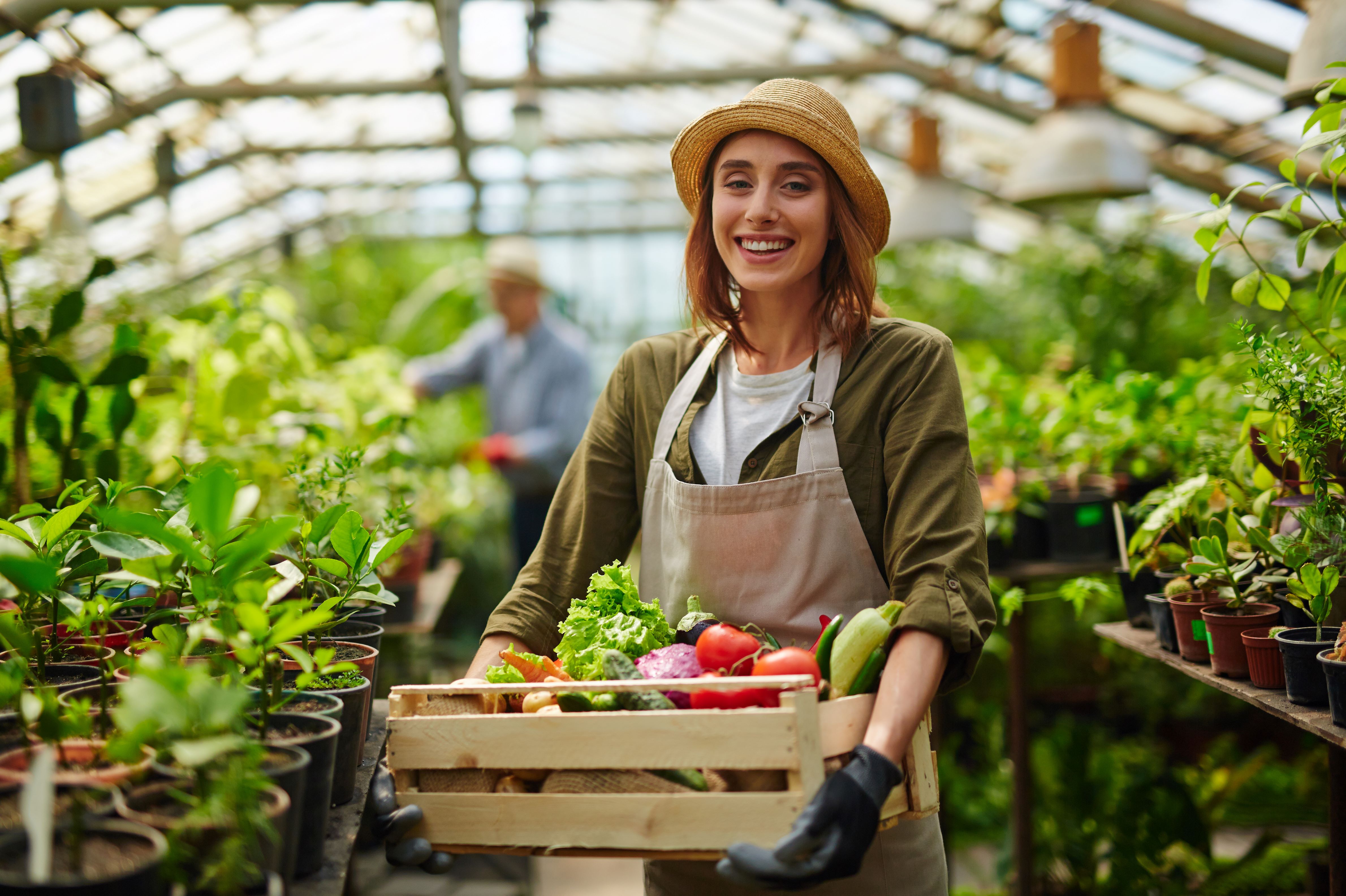 Farmer standing in greenhouse, carrying food items, and surrounded by plants